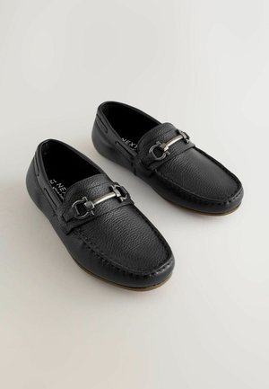 Fekete bőr mokaszinok, texturált felülettel, ezüstszínű fém díszítésekkel és varrott részletekkel a széleken. Kényelmes fel- és levételéhez csúszásmentes kialakítással.