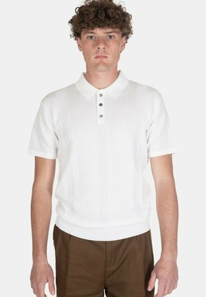 Giovane uomo con capelli ricci che indossa una polo bianca a maniche corte in maglia e pantaloni marroni, in piedi contro uno sfondo chiaro e uniforme.