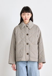 Giacca imbottita color beige con motivo a quadretti, dotata di un grande colletto, chiusura con bottoni e due tasche frontali. Indossata su pantaloni in denim azzurro chiaro.