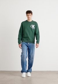 Sweatshirt verde cu guler rotund, având un logo alb pe pieptul din stânga, asortat cu blugi albaștri deschiși și adidași albi, pe un fundal neutru.