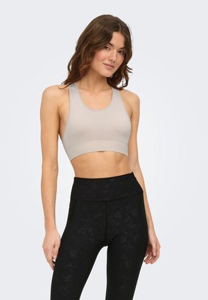 Woman wearing a beige sports bra and high-waisted black patterned leggings, standing with relaxed posture against light background.