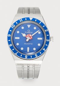 Montre quartz Timex Q avec un cadran bleu, un boîtier argenté et un bracelet en maille. Présente un design de champignon rouge sur le cadran et une lunette bleue avec des indices blancs.