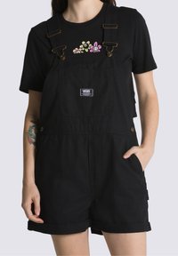 Person wearing black Vans short overalls with front pocket and black t-shirt featuring three colorful smiling flowers.