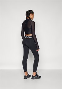 Černý sportovní top s dlouhými průhlednými rukávy, kombinovaný s černými legínami s vysokým pasem, které mají výrazné logo "Nike". Černé sportovní boty.