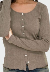 Smeđa rebrasta dugorajna cardigan jakna sa izrezom na vratu i pet bijelih dugmadi sprijeda, s pomalo ispucalim rubom.