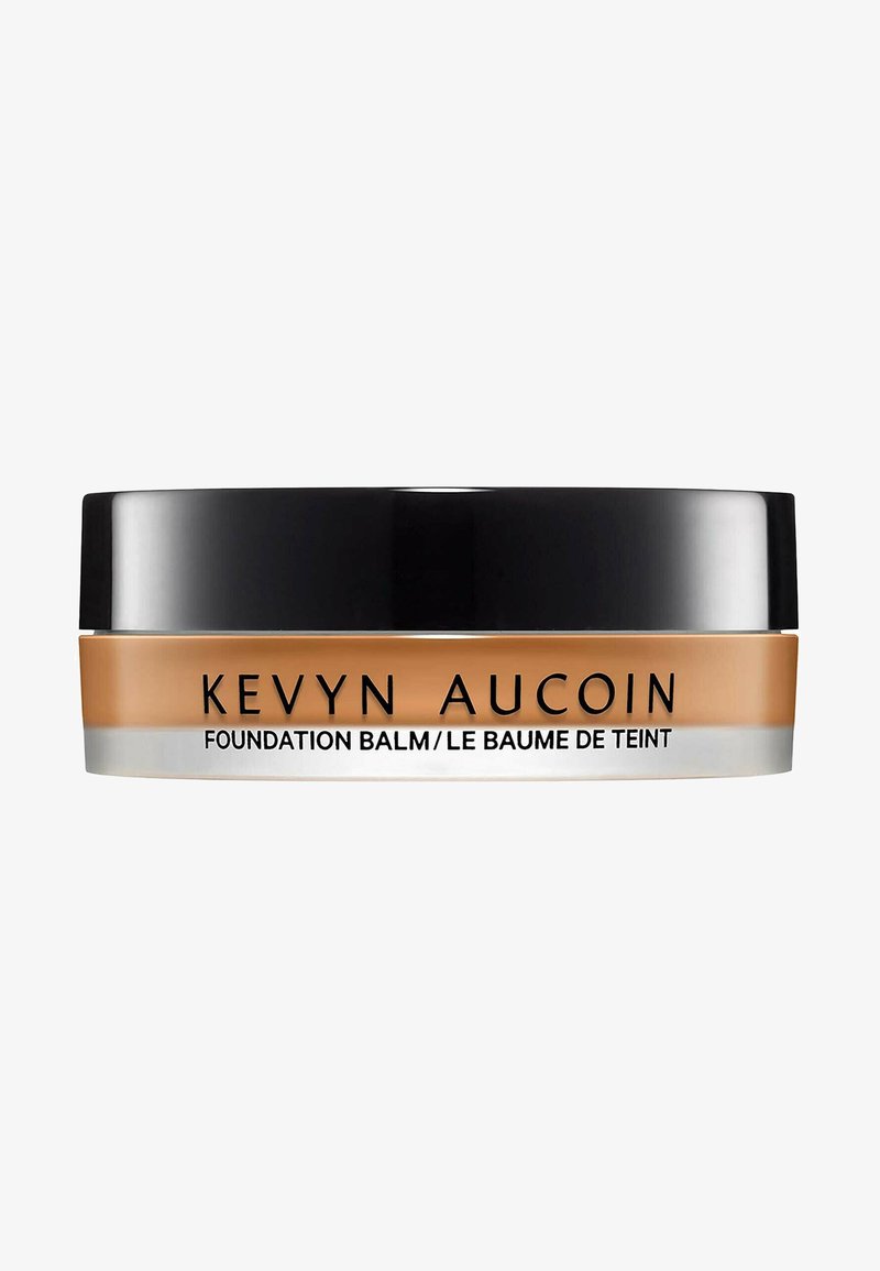 Baume de la Fondation Kevyn Aucoin dans un contenant rond et transparent avec un couvercle noir. Texture lisse et crémeuse dans une teinte beige chaud. Détails en bande argentée.
