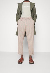 Osoba v zeleném kabátu, světle béžovém tričku s potiskem, béžových kostkovaných kalhotách a hnědých sandálech se dvěma pásky stojí s rukama v kapsách.