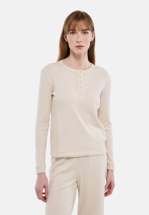 Femme aux cheveux bruns clairs et raides porte un haut beige à manches longues avec des boutons et un pantalon assorti, debout devant un fond clair uni.