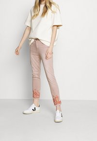 Jeans de algodón rosa con intrincados bordados de paisley naranja en el dobladillo, combinados con una camiseta de corte holgado en crema y zapatillas blancas.