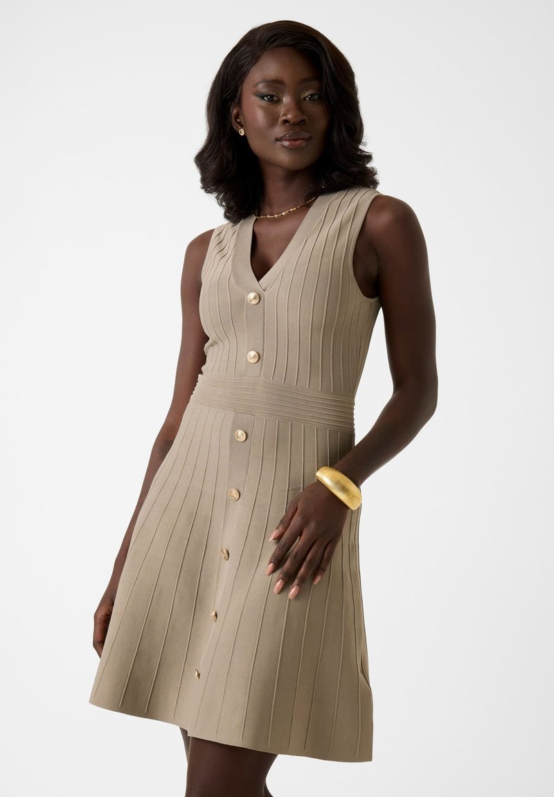 Femme portant une robe beige côtelée sans manches avec des boutons sur le devant et un bracelet en or, posant devant un fond blanc uni.