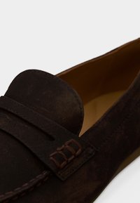 Mocassins en daim marron avec un bout arrondi et des détails cousus. La tige comprend une bride décorative avec des accents et un intérieur lisse.