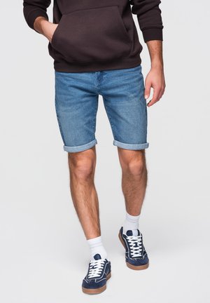 Mies, jolla on käärityt siniset farkkushortsit, valkoiset sukat, tummansiniset tennarit ja tumma huppari, kädet taskuissa.