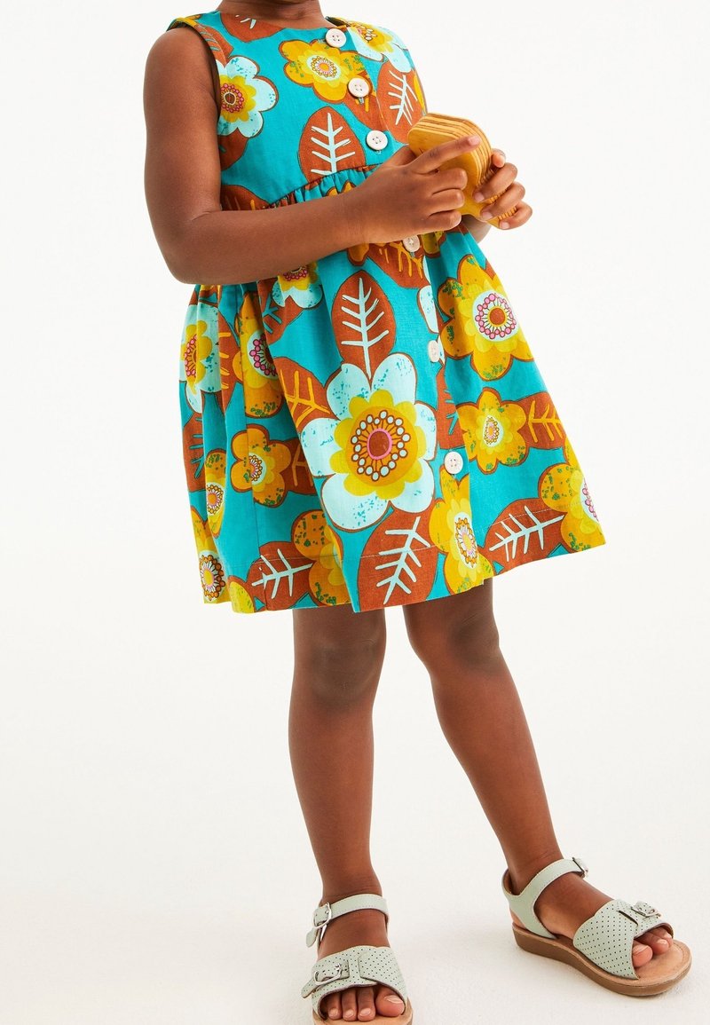 Enfant portant une robe bleue avec de grandes fleurs jaunes et blanches, tenant un petit jouet en bois et portant des sandales de couleur claire.