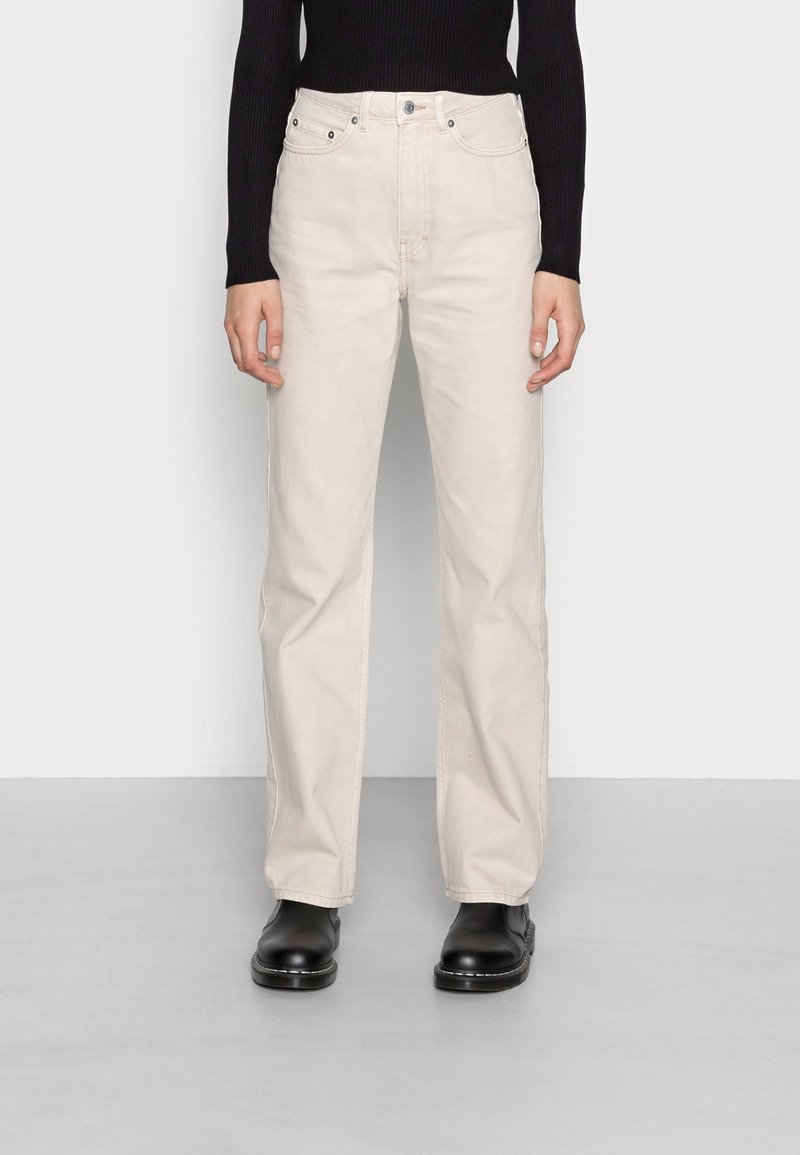 Jeans de pierna ancha beige de algodón, con bolsillos delanteros, trabillas para cinturón y cierre de botón, combinados con botas negras hasta el tobillo.
