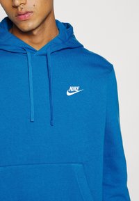 Ung person som bär en ljusblå Nike-hoodie med dragsnören och en framficka, med en del av ansiktet och nacken synlig.