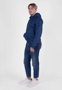 Giacca a vento blu navy con cappuccio e tasche con zip, texture liscia, abbinata a jeans blu sbiaditi e sneakers bianche. Posizione in piedi con le mani in tasca.