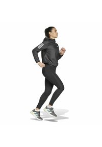 Giacca da corsa nera con tre strisce bianche sulle maniche, leggings neri aderenti e scarpe da corsa grigie con accenti menta, in movimento.