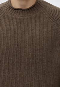 Gros plan d'une personne portant un pull en maille côtelée marron avec un col rond et un tissu texturé.