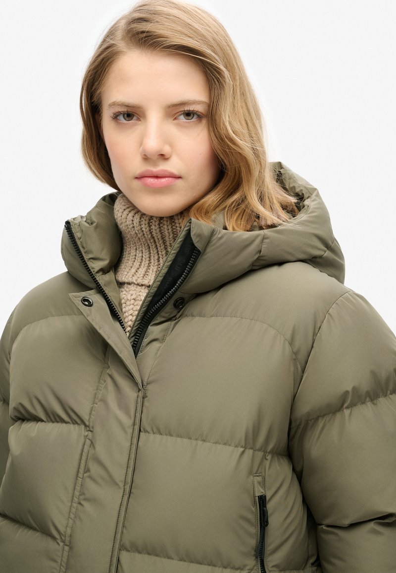 Olivgrüne Steppjacke mit Kapuze, mit quilted Bereichen, einem Reißverschluss und Druckknöpfen, getragen über einem beigen Strickpullover.