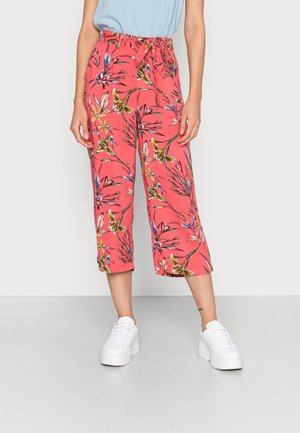 Femme portant un pantalon court corail avec un imprimé floral multicolore et des baskets blanches à plateforme, debout devant un fond uni.
