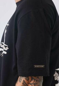 Zwart T-shirt met ronde hals en korte mouwen, voorzien van een grafische print en een klein metallic label op de mouw met de tekst "PROJECT X PARIS."