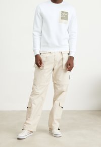Sudadera blanca de cuello redondo con un parche de logo beige, combinada con pantalones cargo beige que tienen bolsillos laterales. Se lleva con zapatos de tela de cuadros blancos.