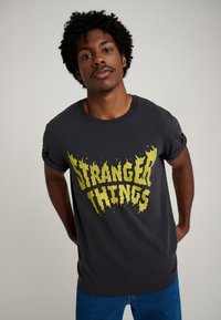 Tummanharmaa puuvillainen t-paita, jossa on rohkea keltainen "STRANGER THINGS" -grafiikka liekinomaisella fontilla. Rentoa istuvuutta ja käärityt lyhyet hihat.