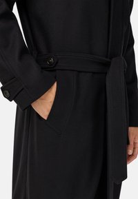 Rokas kabatā melnā mētelī ar jostu, pogām un sānu cilpām neitrālā fonā.