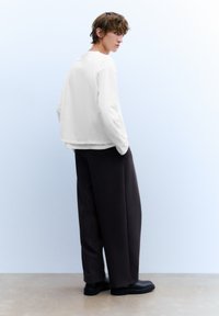 Weißes Langarmhemd mit rundem Ausschnitt, kombiniert mit locker sitzender dunkler Hose und schwarzen Schuhen, stehend vor einem blassblauen Hintergrund.