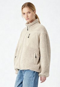 Giovane donna con capelli biondi indossa una giacca beige in pile con zip e jeans blu, in piedi davanti a uno sfondo bianco semplice, rivolta verso l'obiettivo.