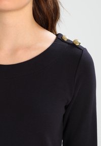 Camiseta negra de manga larga hecha de tejido de punto, con un escote redondeado y tres botones decorativos dorados en el hombro izquierdo.