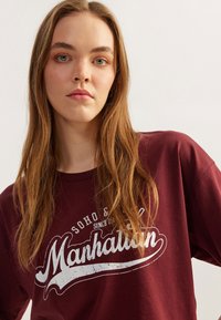Bordowa bawełniana koszulka z krótkim rękawem z białym, zniszczonym nadrukiem graficznym "Manhattan". Luźny krój. Gładka tekstura.