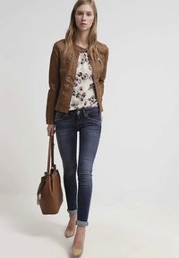 Junge Frau läuft, trägt eine braune Lederjacke, eine blumige Bluse, dunkle Skinny-Jeans, beige Absatzschuhe und trägt eine große braune Handtasche.