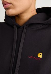 Black hoodie made of cotton blend, featuring a small embroidered logo in yellow and red. Drawstrings and soft texture visible.