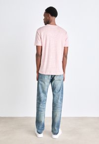 Gaiši rozā īsām piedurknēm t-krekls, izgatavots no mīkstas auduma. Valkāts ar zili izbalinātiem džinsiem un baltiem sporta apaviem. Skats no aizmugures.