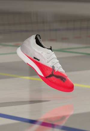 Λευκό και κόκκινο ποδοσφαιρικό παπούτσι Puma Nitro με κορδόνια να αιωρείται πάνω από το δάπεδο ενός γυμναστηρίου με έγχρωμες γραμμές.