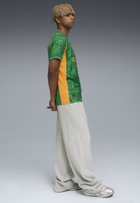 Grön och gul sporttröja med ett texturerat mönster, i kombination med ljusgrå oversizedbyxor och flerfärgade sneakers, sedd från sidan.