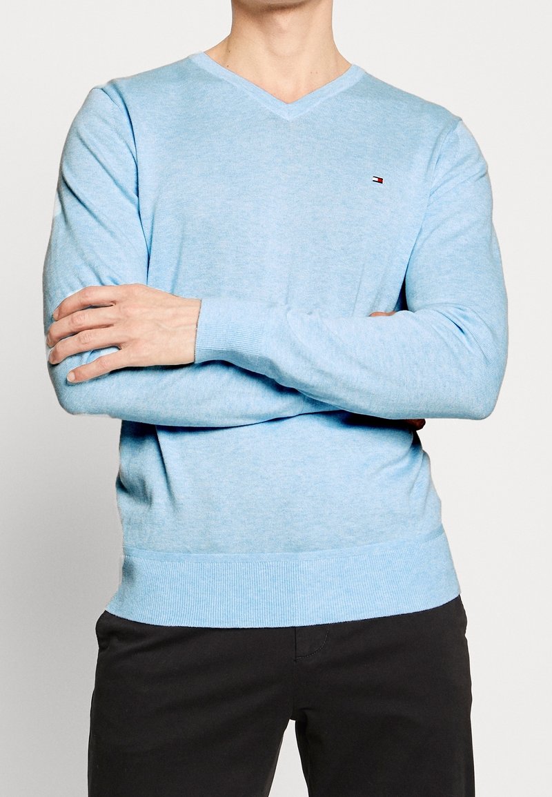 Helle blaue V-Ausschnitt-Strickjacke aus weichem Material, mit gerippten Bündchen und Saum, und einem kleinen Logo auf der Brust. Trägt dazu dunkle Hose.