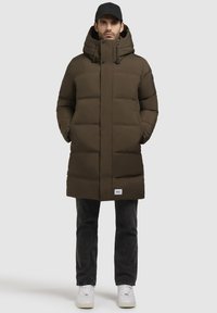 Braune Puffjacke mit hohem Kragen, verstellbarem Kapuzenzug und Knopfdetails. Getragen über dunklen Jeans und weißen Sneakern.