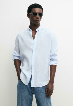 Man die een lichtblauw overhemd met knopen draagt met opgerolde mouwen, een blauwe spijkerbroek en ronde donkere zonnebril, staat met één hand in de zak.