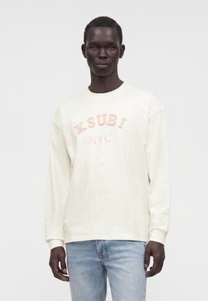 Homme grand portant une chemise blanche à manches longues avec "KSUBI NYC" en lettres rose clair et un jean bleu clair éclaboussé de peinture sur un fond uni.