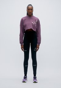 Μωβ cropped φούτερ με εκτύπωση, σε συνδυασμό με μαύρο κολάν με ανακλαστικές ρίγες και μωβ αθλητικά παπούτσια.