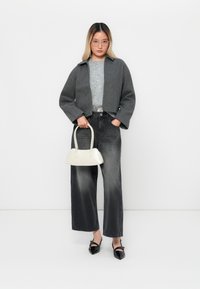 Giacca grigia corta, maglione grigio chiaro, jeans neri a vita alta, scarpe con tacco nere e una piccola borsa bianco sporco. Sfondo neutro.