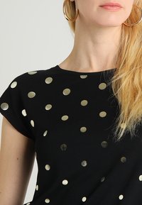 Svart t-shirt med metalliska guldprickar, korta ärmar och rund halsringning. Slätt tyg med en avslappnad passform.