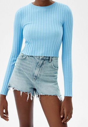 Kvinde iført en lyseblå ribstrikket crop top med lange ærmer og højtaljede distressed denimshorts mod en hvid baggrund.