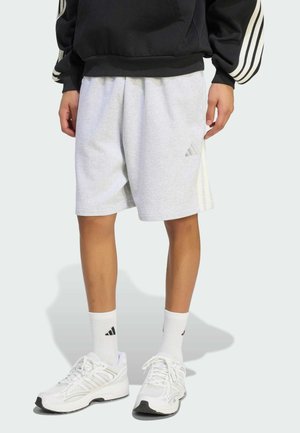 Persoon draagt lichtgrijze Adidas-short, witte Adidas-sokken en witte sneakers, staand tegen een effen lichte achtergrond.