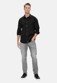 Camicia nera a maniche lunghe con bottoni, indossata con jeans grigi e sneakers nere. La camicia presenta un logo discreto sul petto e polsini arrotolati.