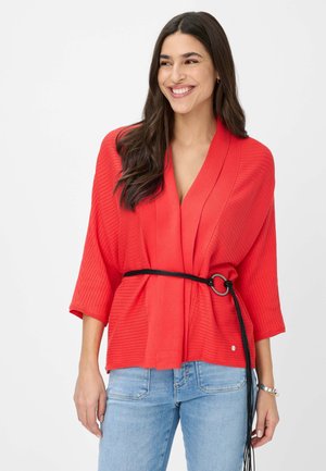 Haut enveloppant rouge à côtes avec de larges manches et un décolleté en V profond, cintré à la taille avec une ceinture noire, associé à un jean bleu.