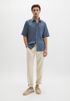 Ein Mann steht mit den Händen in den Taschen, trägt ein blaues kurzärmliges Hemd mit Knöpfen, beige hochgekrempelte Hosen und beigefarbene Turnschuhe vor weißem Hintergrund.