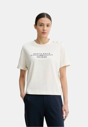 Kobieta o ciemnych włosach w białym t-shircie North Sails Guess z guzikami na ramieniu, w ciemnych spodniach, stojąca z jedną ręką w kieszeni.